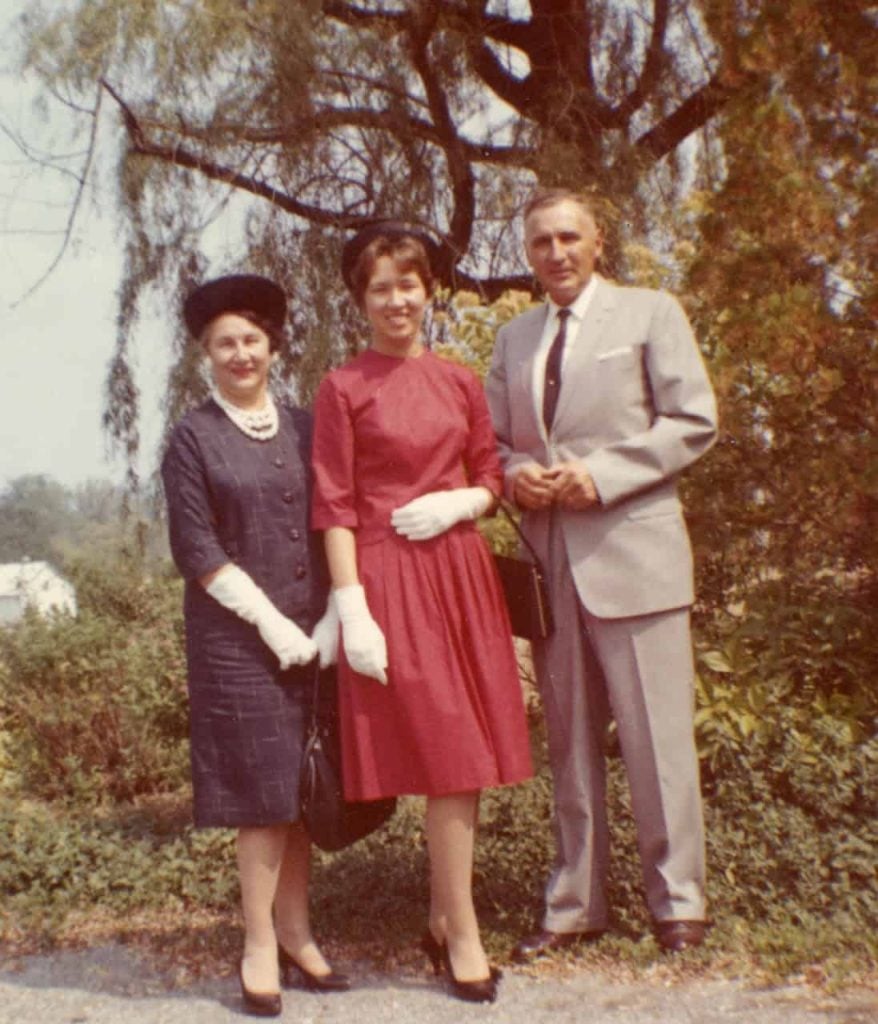 A photo of a man and woman with their young daughter, maybe 20 years old, in between them all smiling outdoors standing in front of a tree. They are wearing formal attire, the man in a grey suit, mother in a black dress and gloves and a black hat and the daughter wearing a red dress, white gloves and black hat and heels.