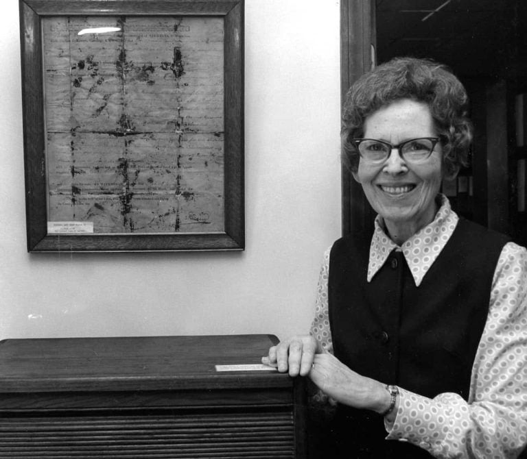 Archival black and white photo of a woman in glasses standing next to a framed historical document.