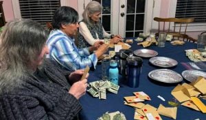 Three older women, two with long grey hair and one with shorter black hair are sitting at a table counting donations collected by students. The donation envelopes are decorated with hearts.