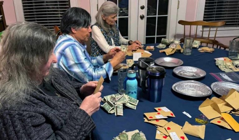 Three older women, two with long grey hair and one with shorter black hair are sitting at a table counting donations collected by students. The donation envelopes are decorated with hearts.