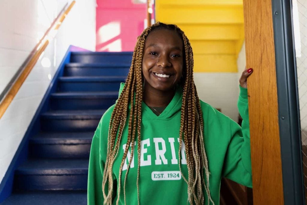 A young black woman with her hair in box braids down to her waist stands smiling at the bottom of blue painted steps. She is wearing a bright green sweatshirt that says "Nerinx, est. 1924)