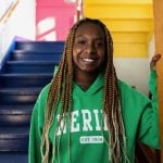 A young black woman with her hair in box braids down to her waist stands smiling at the bottom of blue painted steps. She is wearing a bright green sweatshirt that says "Nerinx, est. 1924)