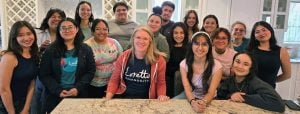 A large group 16 Latino college students of varying ages and one white woman in her 40s smiling together at an end of year retreat.