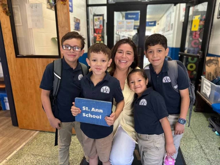 Four elementary school students with blue polo shirts and khakis taking a picture with a teacher who is squatted down to be the student's height. One of the students is carrying a blue plaque that says "St. Ann School"
