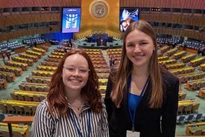 Two young college aged students smilng together in front of the UN general assembly hall. Behind them is an impressive room with hundreds of seats and the UN emblem centered over the podium with two large screens on each side.