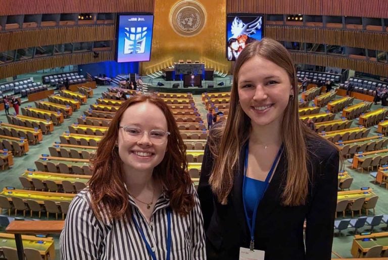 Two young college aged students smilng together in front of the UN general assembly hall. Behind them is an impressive room with hundreds of seats and the UN emblem centered over the podium with two large screens on each side.