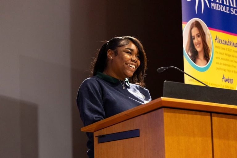 A young Black woman smiling brightly as she stands and talks at a podium. She is wearing a blue sweatshirt and a silver necklace with her hair brown shoulder length hair straightened and pulled back with a clip on one side.