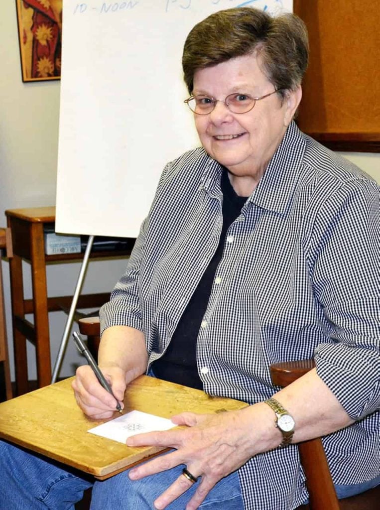 An older woman with short brown hair, small round glasses and a checkered shirt smiles and looks up from drawing a mandala.