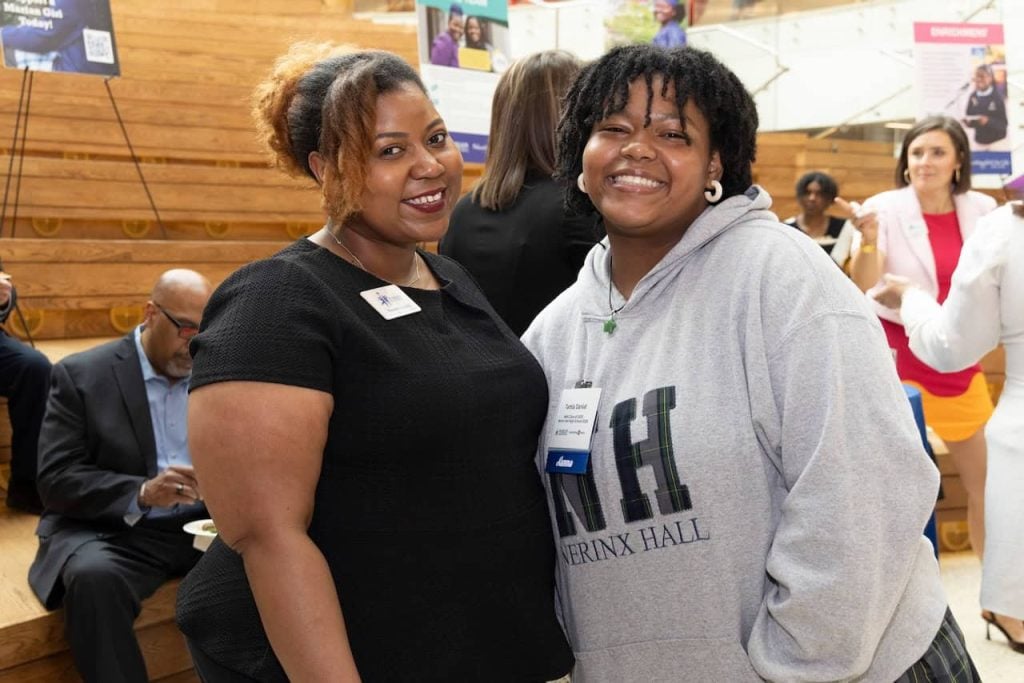 A Black woman with her hair up and a black shirt smiles next two a Black high school girl who has shorter hair in twists and is wearing a grey Nerinx Hall sweatshirt.