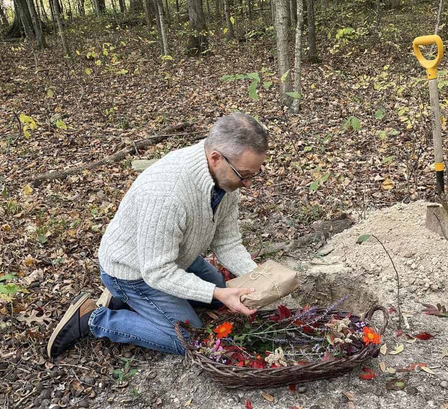 A man in a quarter-zip sweater and jeans kneels on the ground in the woods. He holds a package wrapped in brown paper and twine in one hand that he has removed from a basket full of flowers and is placing into the ground. A shovel sticking out of a pile of dirt is visible on the right side of the photo.