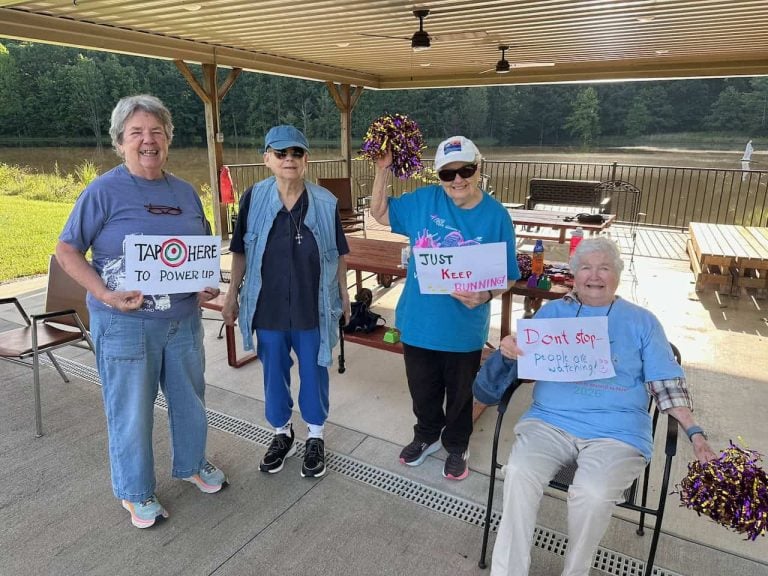 Four elder women standing and sitting under an awning holding posters on a 5k race for the runners. The posters say "Dont stop people are watching" "Just keep running" and "tap here to power up"