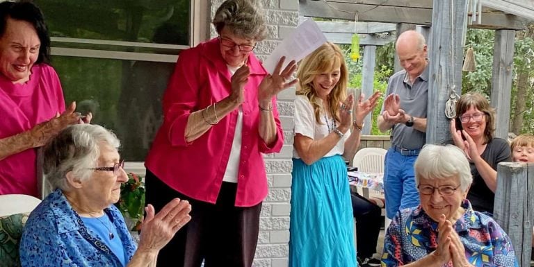 Two women turning 90 are shown smiling and being celebrated by people all around them at a party clapping and smiling