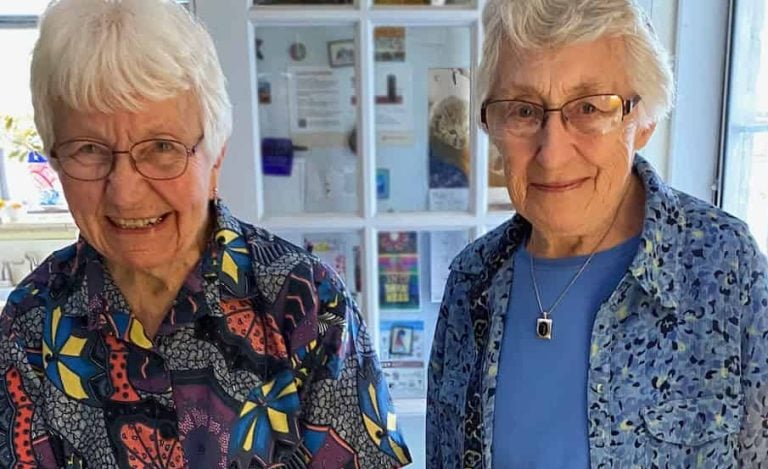 Two women in colorful blouses and short white hair and glasses smile together in front of a yellow cake that says "Happy 90th Birthday Joan and Ellen!"