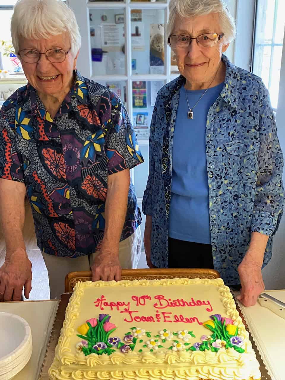 Two women in colorful blouses and short white hair and glasses smile together in front of a yellow cake that says "Happy 90th Birthday Joan and Ellen!"