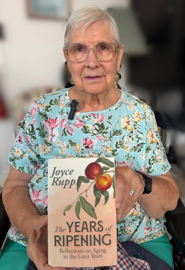 An older woman wearing a flowered shirt with a microphone attached to the collar holds up a book by Joyce Rupp entitled "The Years of Ripening; Reflections on Aging in the Later Years."
