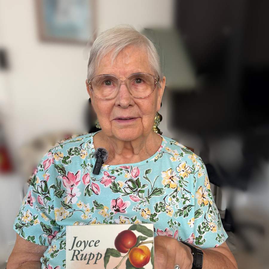 An older woman wearing a flowered shirt with a microphone attached to the collar holds up a book by Joyce Rupp.