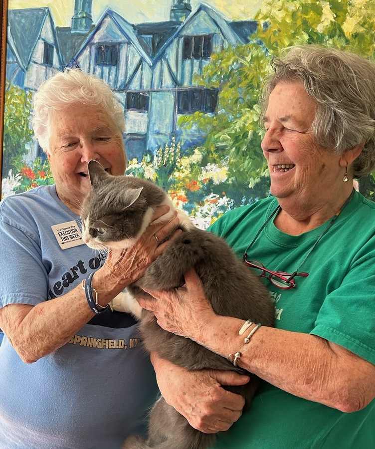 Two older women lovingly cuddle a large, fluffy gray-and-white cat.