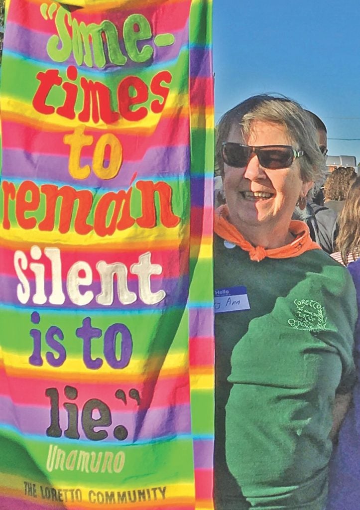 An older white woman with short gray hair, sunglasses and a green t-shirt holds a colorful rainbow banner that says "sometimes to remain silent is to lie."