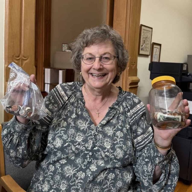 A woman with a huge smile stands holding a bag of coins in her right hand and a jar of coins and bills in her left.