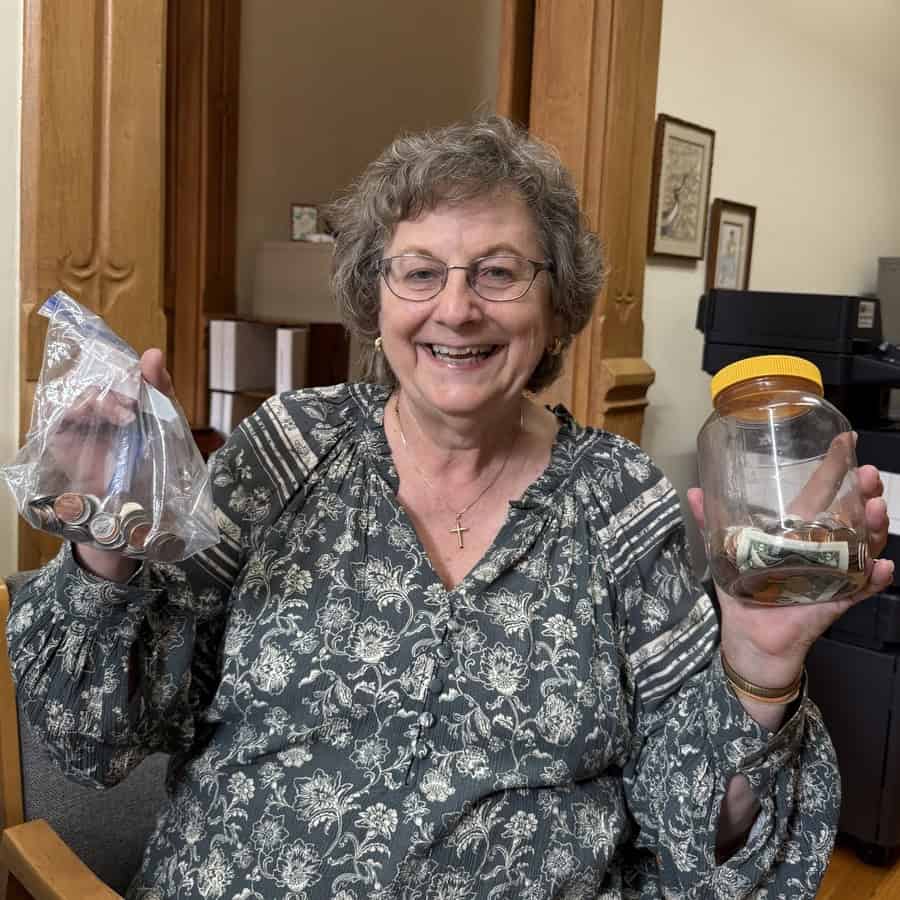 A woman with a huge smile stands holding a bag of coins in her right hand and a jar of coins and bills in her left.