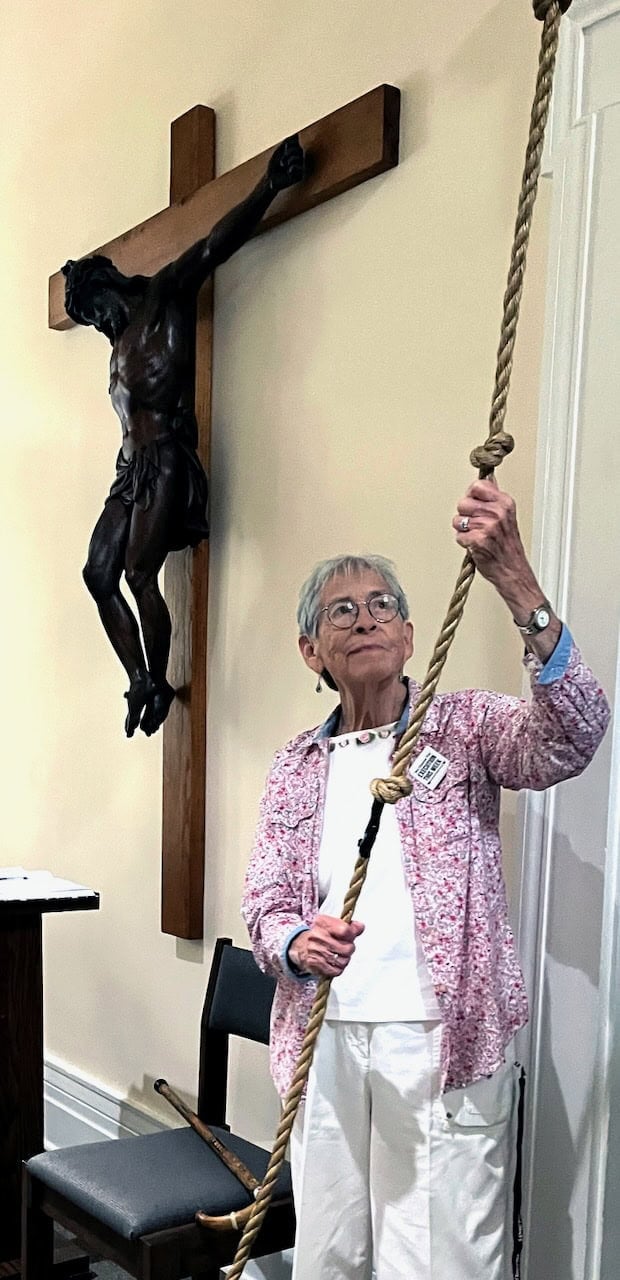 An older religious sister with short grey hair, glasses, a white shirt and pants and a purple patterned cardigan holding a rope to toll a church bell.