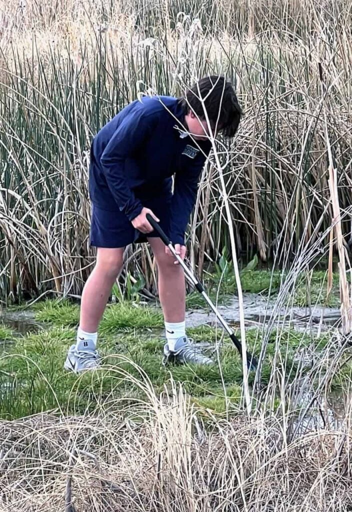 An elementary school boy with short brown hair, a blue long sleeve shirt and blue shorts with grey sneakers is shown picking up trash outside.