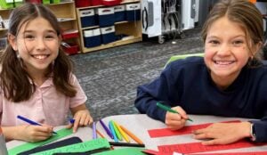 Two elementary aged girls take a break to smile for a picture as they are holding markers andd making placemats and paper floral centerpieces for an upcoming event.
