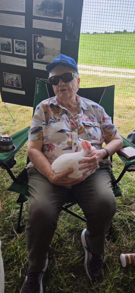 An older woman in a floral shirt, dark green pants, sneakers, a blue hat and sunglasses sitting in a green lawn chair outside holding a white bunny rabbit.