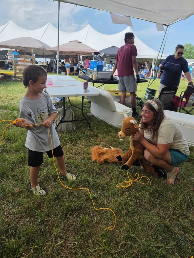 A little kid practices lassoing a stuffed animal held by an an adult squatting on the ground. They are both laughing and smiling.