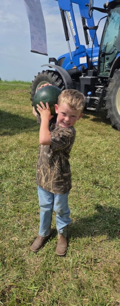 A small little boy in a camo shirt, jeans and brown boots proudly holding up a watermelon in front of a tractor.