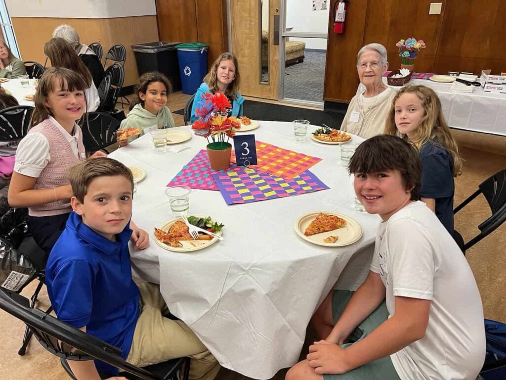 A religious sister with short white hair, glasses and a cream shirt sits with six elementary aged students for lunch.