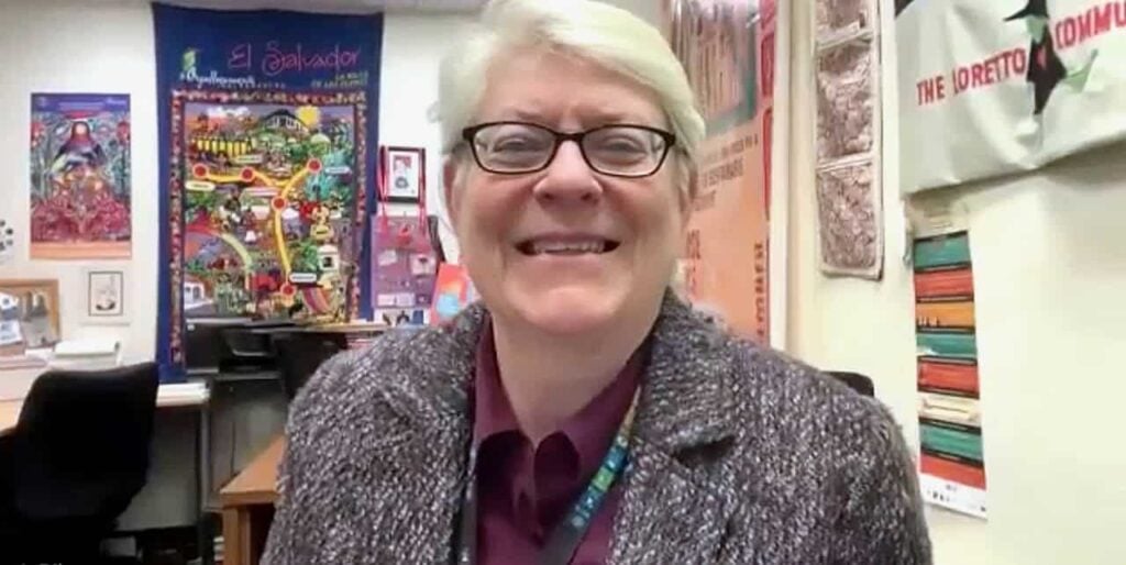 A middle aged white woman smiling with short white hair, black glasses, a maroon collared shirt and a tweed jacket in an office full of colorful posters and banners behind her.