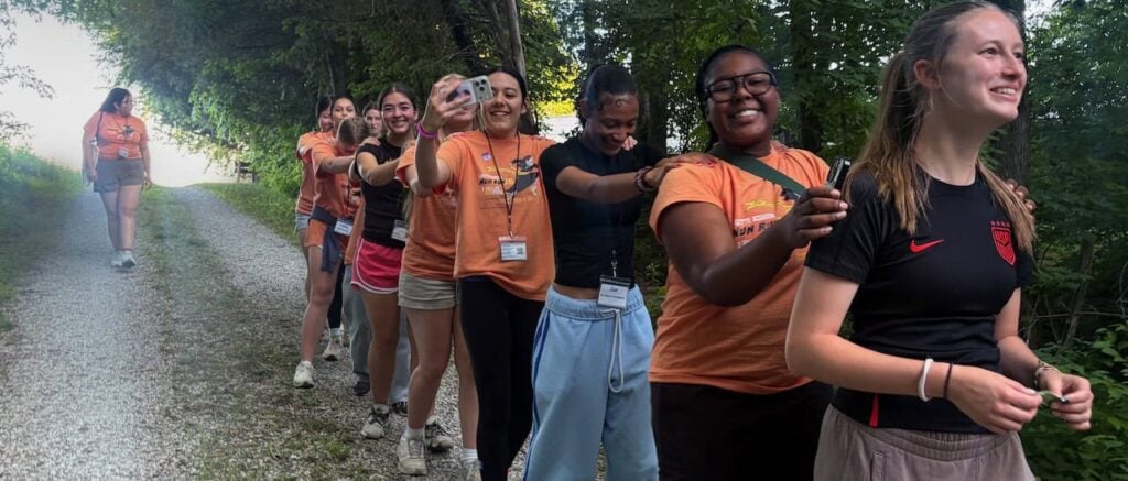 A group of 10 high school girls in mostly orange shirts hands on each others shoulders in a line laughing and smiling on a gravel path in the woods.