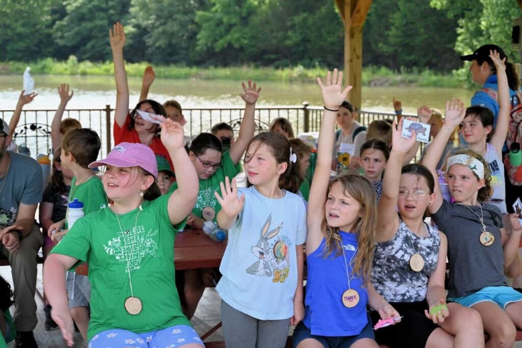 A group of children sitting outside at a day camp all raising their hands and engaging the lesson enthusiastically.