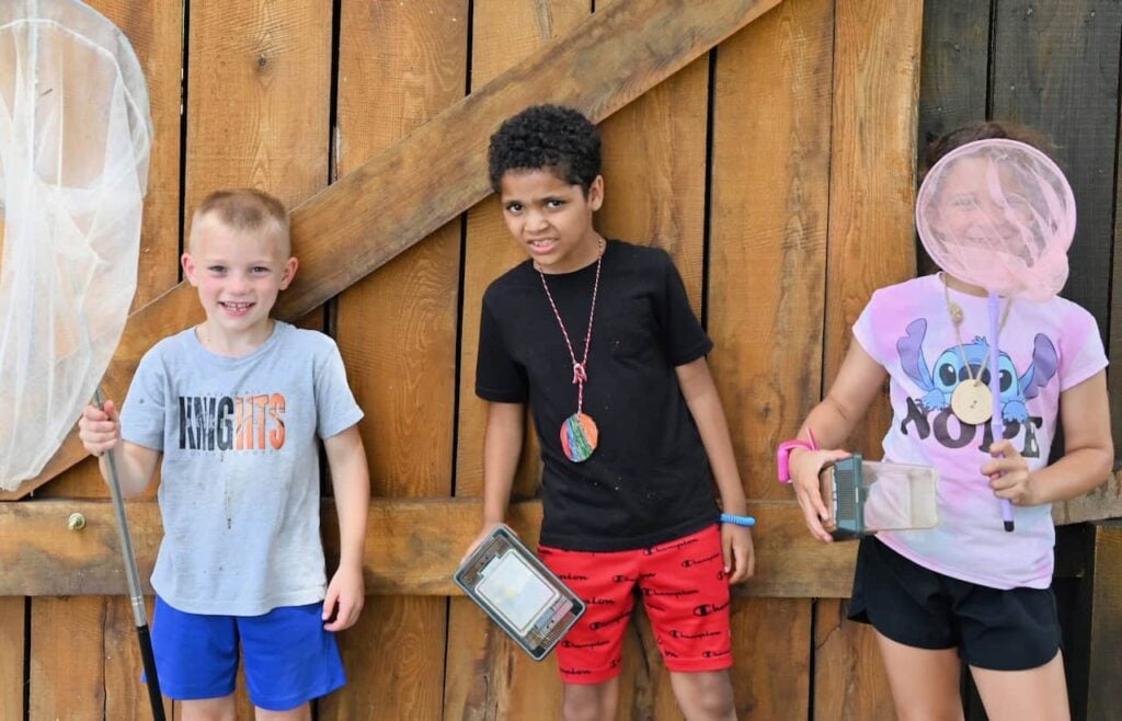 Three elementary school kids at a day camp pause and smile for a photo shoot. Two of the campers have butterfly nets.