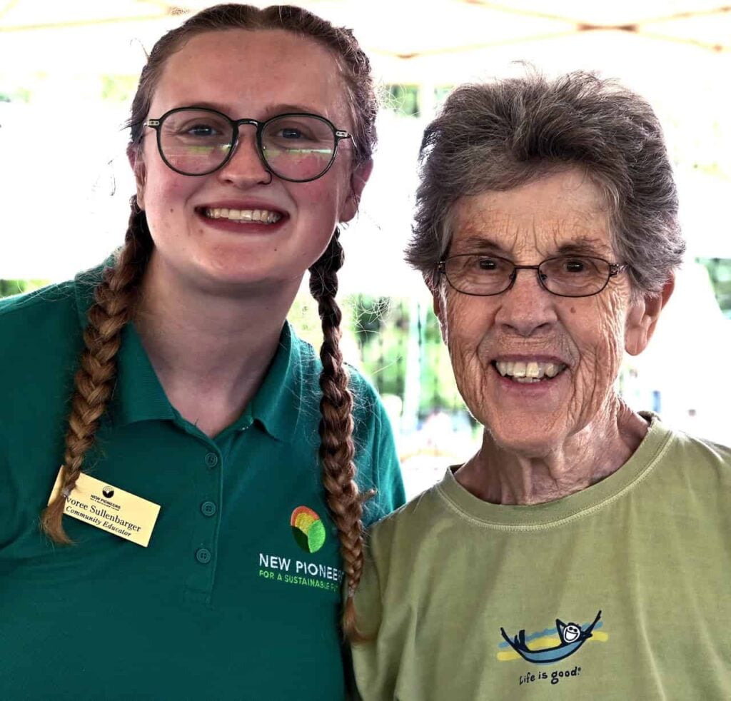 A younger and older woman take a bright and smiley photo together outdoors at an agricultural event. The younger woman on the left has her hair in two braids and is wearing a dark green polo shirt. The older woman on the right has short hair, and is wearing a light green "life is good" shirt. They are both wearing black framed glasses.