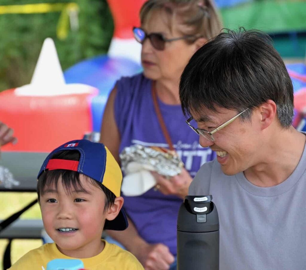 A father and son at an outdoor event. The father smiles widely at his son as his son looks onward in awe at something. The kid is wearing a backwards baseball cap and holding a water bottle. His dad is wearing a grey t-shirt and glasses and holding a black water bottle.