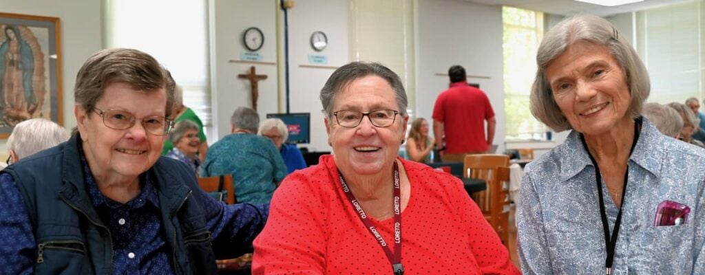 Three elder women smile for a group photo at a gathering. The woman on the left has brown hair, glasses, a blue long sleeved shirt and blue vest. The woman in the middle has short grey hair, glasses, and polka dot red long sleeved shirt. The woman on the right has a white bob with hair clips pulling the hair out of her face wearing a blue and white collared shirt.