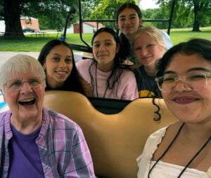 A group of five high school girls smile with a nun on a golf cart outside.