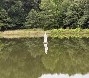 A statue of the Virgin Mary stands in the middle of a lake with the reflection of the woods behind her.