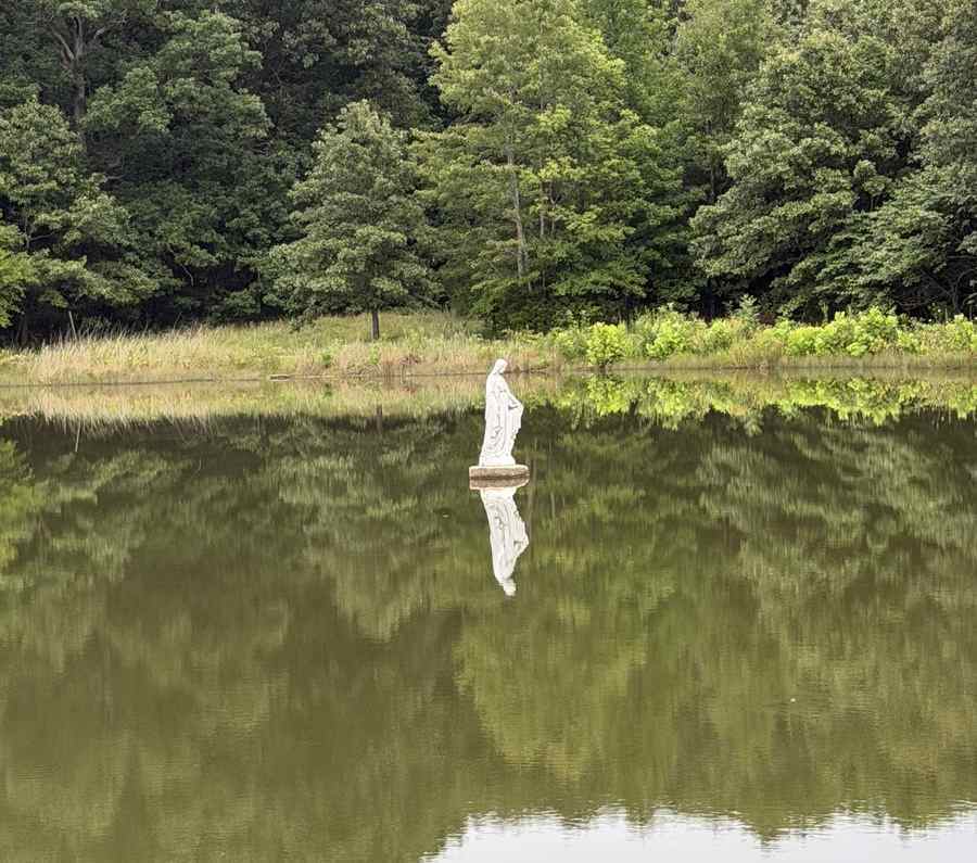 A statue of the Virgin Mary stands in the middle of a lake with the reflection of the woods behind her.