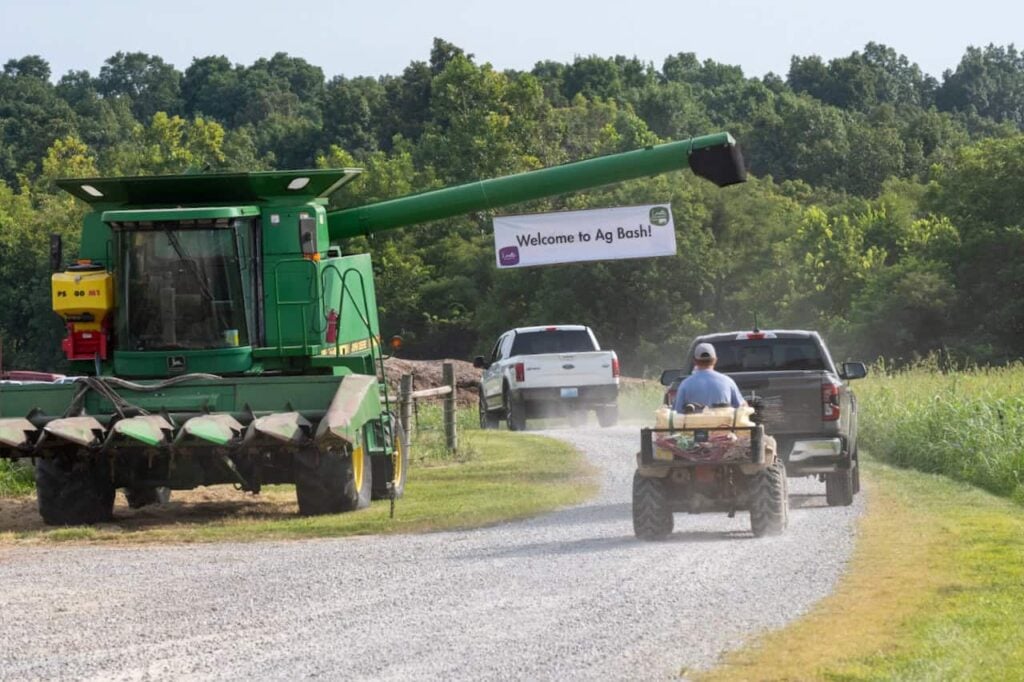 A sign hanging on a tractor that says "Welcome to Ag Bash" that is hanging over a dirt road. Two pickup trucks and a man on a small tractor drive on the road to the Ag Bash.