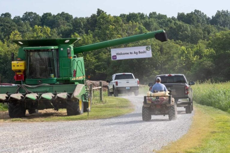 A sign hanging on a tractor that says "Welcome to Ag Bash" that is hanging over a dirt road. Two pickup trucks and a man on a small tractor drive on the road to the Ag Bash.