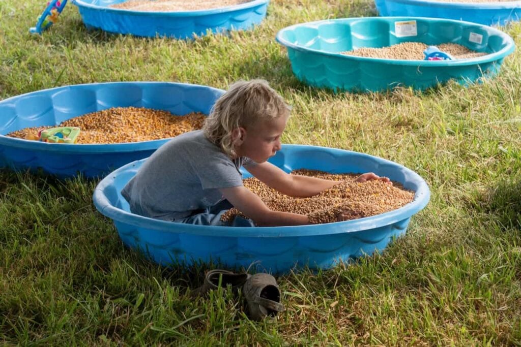 A little kid is pictured sitting in a blue kiddie pool filled with corn or another grain very concentrated on feeling the grain with his arms. His camo crocs sit outside of the kiddie pool.