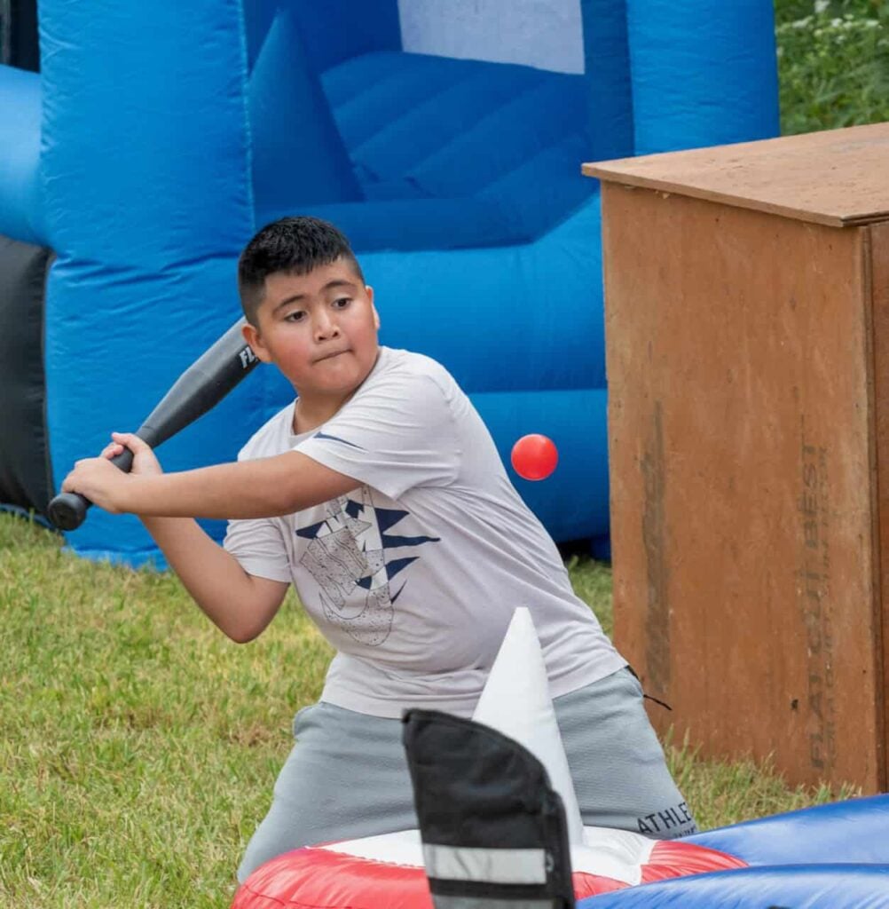 An older elementary aged kid is pictured mid-shot playing baseball and is about to hit a red ball with a black bat.