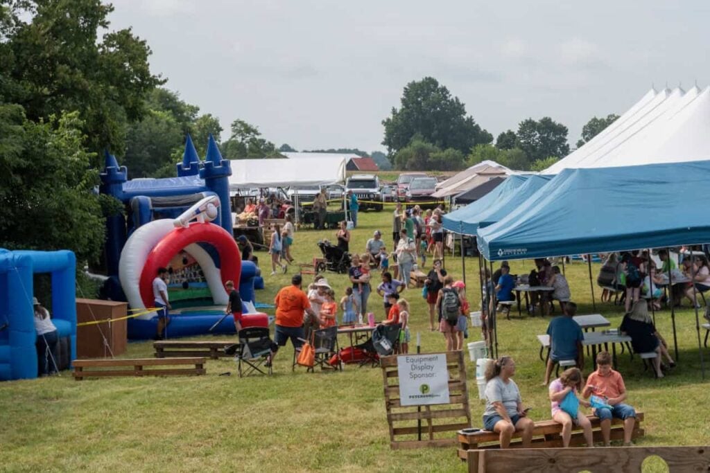 A sunny day on Ag Bash 2026. A wide shot of various tents, blow up bouncy houses and participants standing, walking and sitting at tables and enjoying the day.