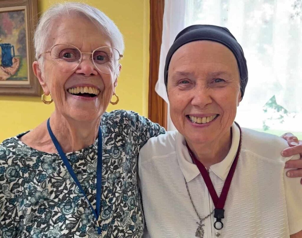 Two elder women smiling and delighting in each other's company. The woman on the left has short white hair, big glasses, gold hoop earrings and a paisley blue shirt. The woman on the right has her hair in a dark head wrap and is wearing a white collared shirt.