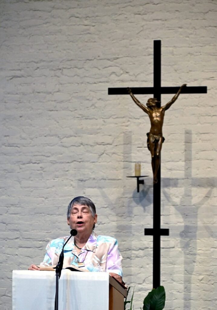 An older woman with brown and grey hair sings joyfully at a podium at Mass. A crucifix and white brick wall are behind her.