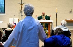 A closeup of the back of an older woman standing up in between older women in wheelchairs as a priest in front of her presides over Mass.