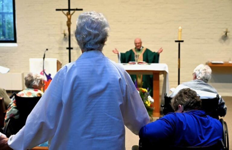 A closeup of the back of an older woman standing up in between older women in wheelchairs as a priest in front of her presides over Mass.